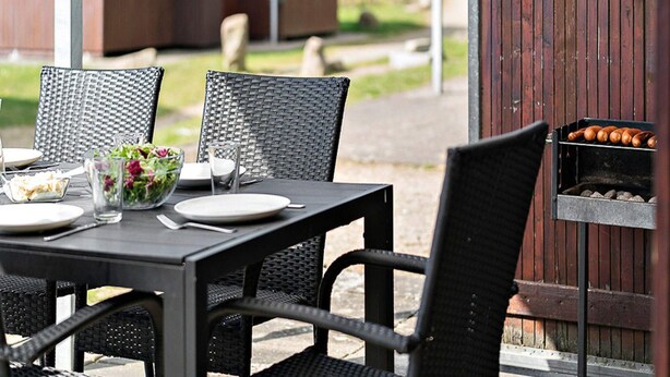 En uteservering med ett bord dukat för två personer, omgiven av flätade stolar. En grill med korv syns i bakgrunden.