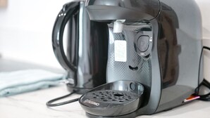 Máquina de café preta com suporte para cápsulas Tassimo, cabo de alimentação e painel de controle com botão e visor.