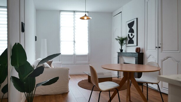 Uma sala de estar moderna com uma mesa redonda de madeira, duas cadeiras, uma planta grande em vaso e um sofá branco.