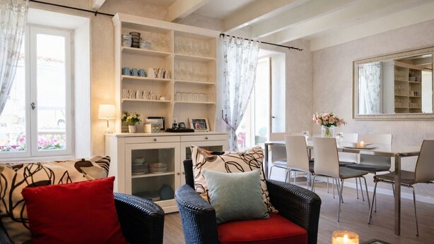 Una sala de estar con sofá, sillón y mesa de comedor. Se observa una estantería con diversos objetos y un espejo sobre la mesa de comedor.