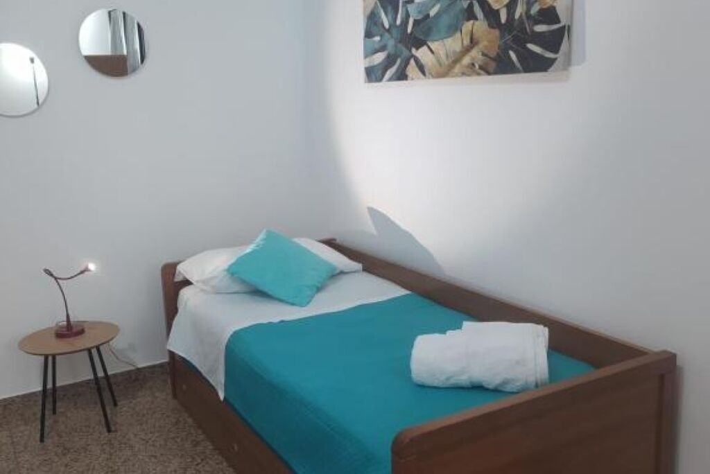Uma cama de solteiro com estrutura de madeira, uma colcha verde-água e um travesseiro branco. Há uma pequena mesa redonda com uma luminária ao lado da cama.