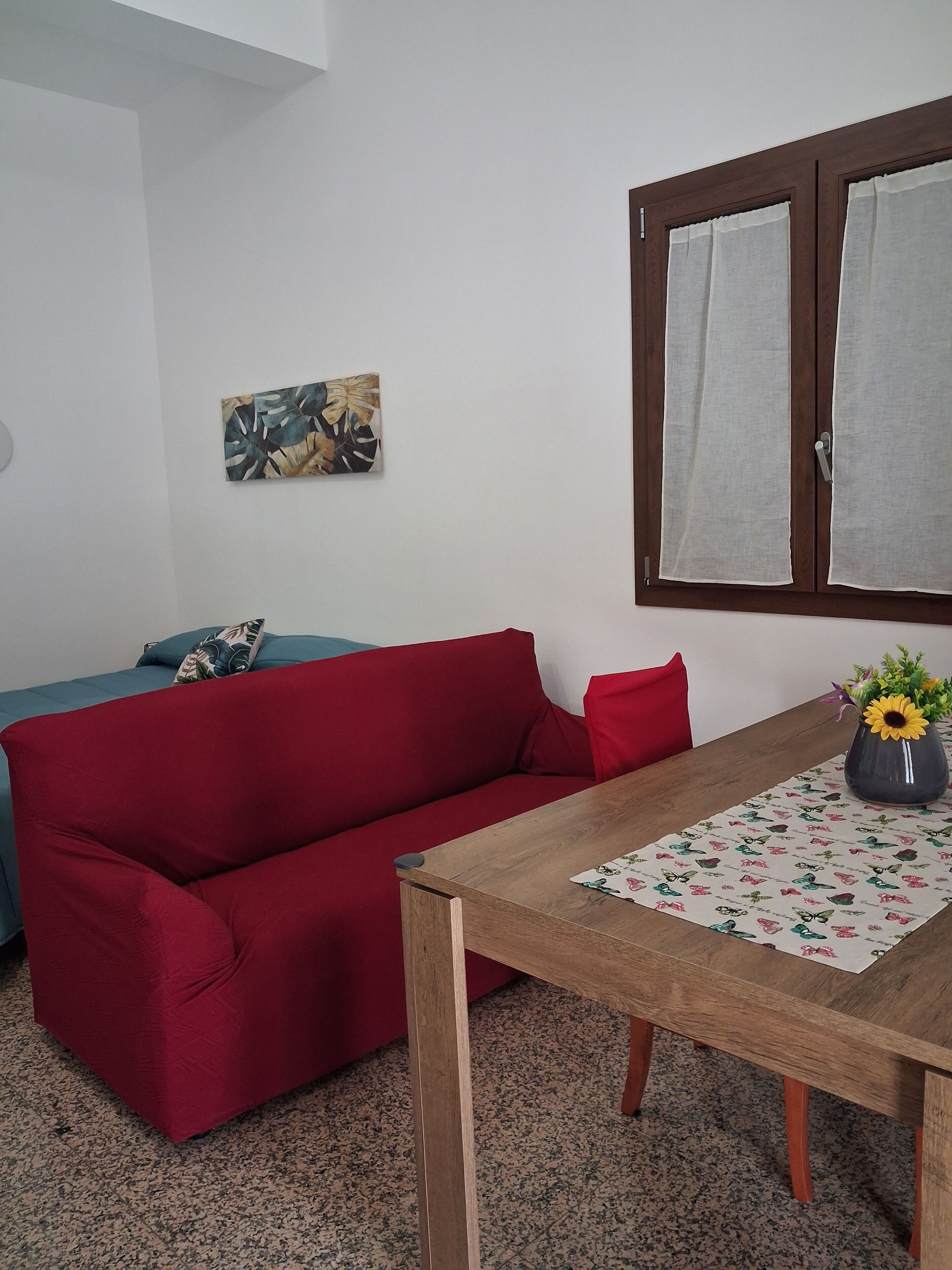 Ein rotes Sofa, ein Holztisch mit einer blumigen Tischdecke, eine Vase mit Blumen und eine Wand mit einem Gemälde.