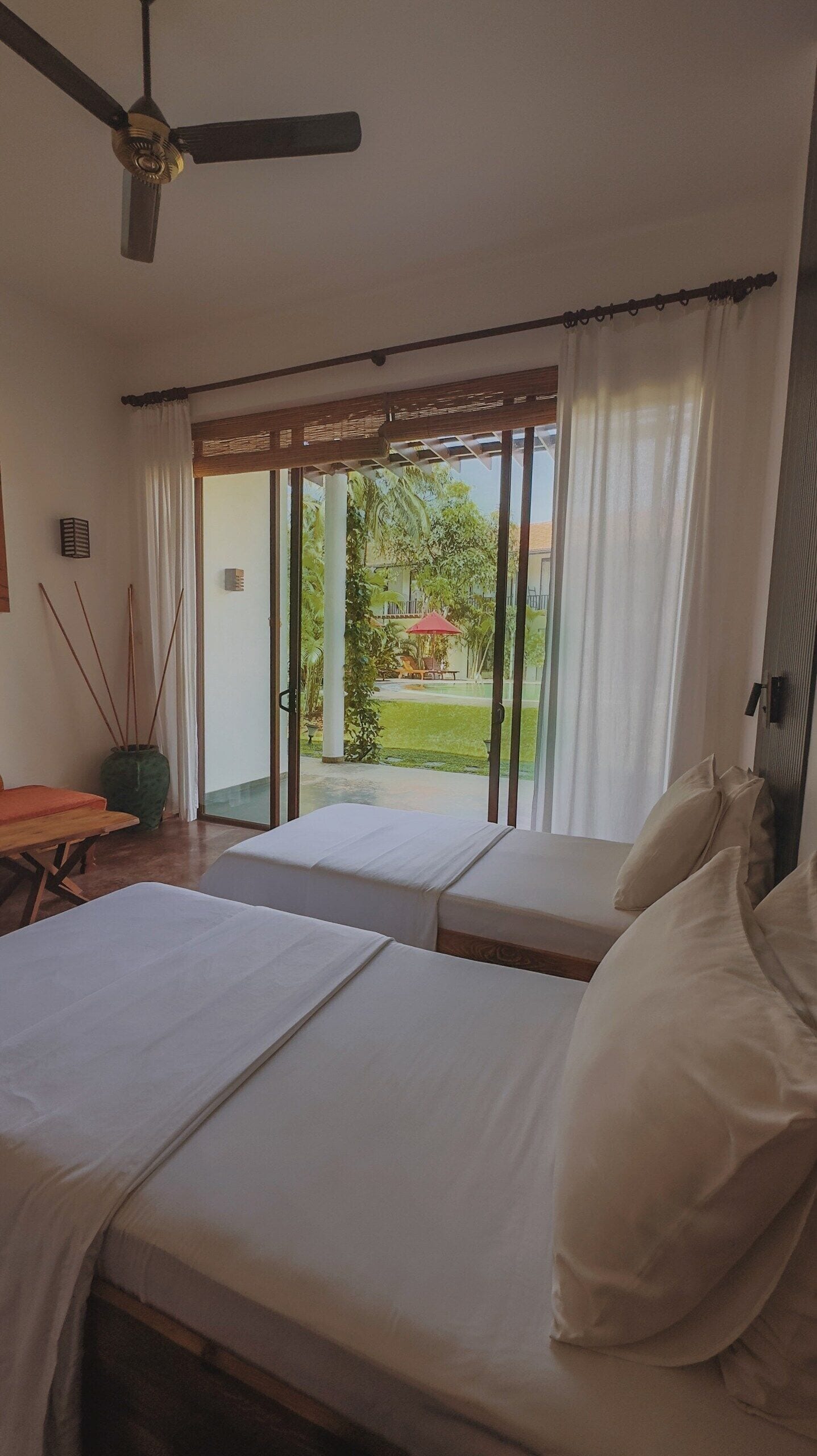 Une chambre avec deux lits, un ventilateur de plafond et une vue sur un jardin, accessible par une porte-fenêtre coulissante.