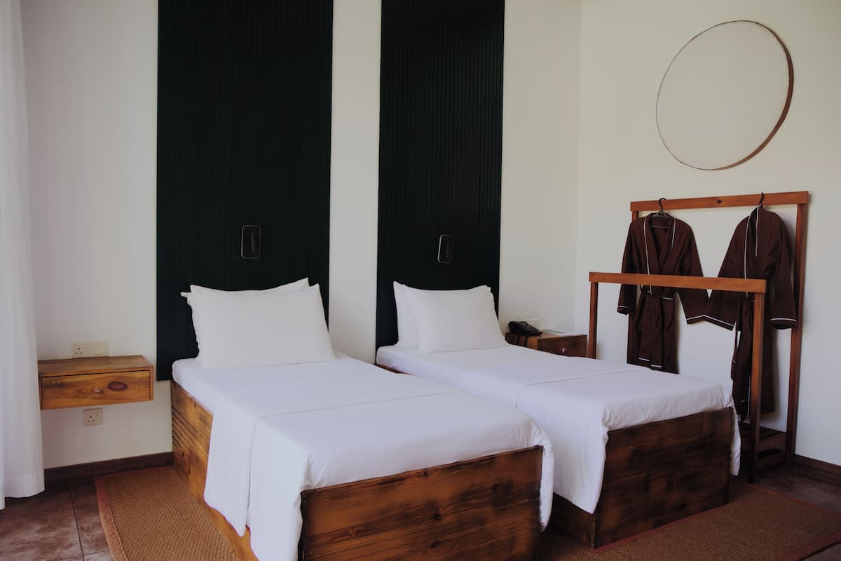 Una cama doble con sábanas blancas, una mesita de noche de madera, un perchero y un espejo circular fijado a la pared.