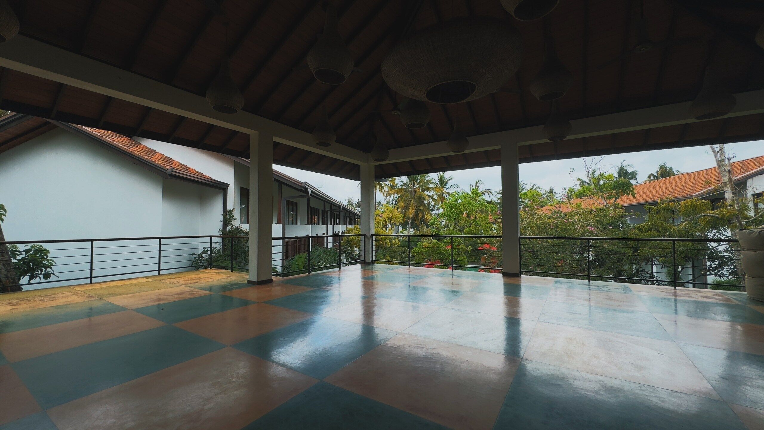 An open covered area with a tiled floor, a white building with a red-tiled roof, and a view of palm trees and other greenery.