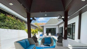 Uma área externa coberta com piscina, dois sofás de vime com almofadas azuis e uma cadeira de vime redonda.
