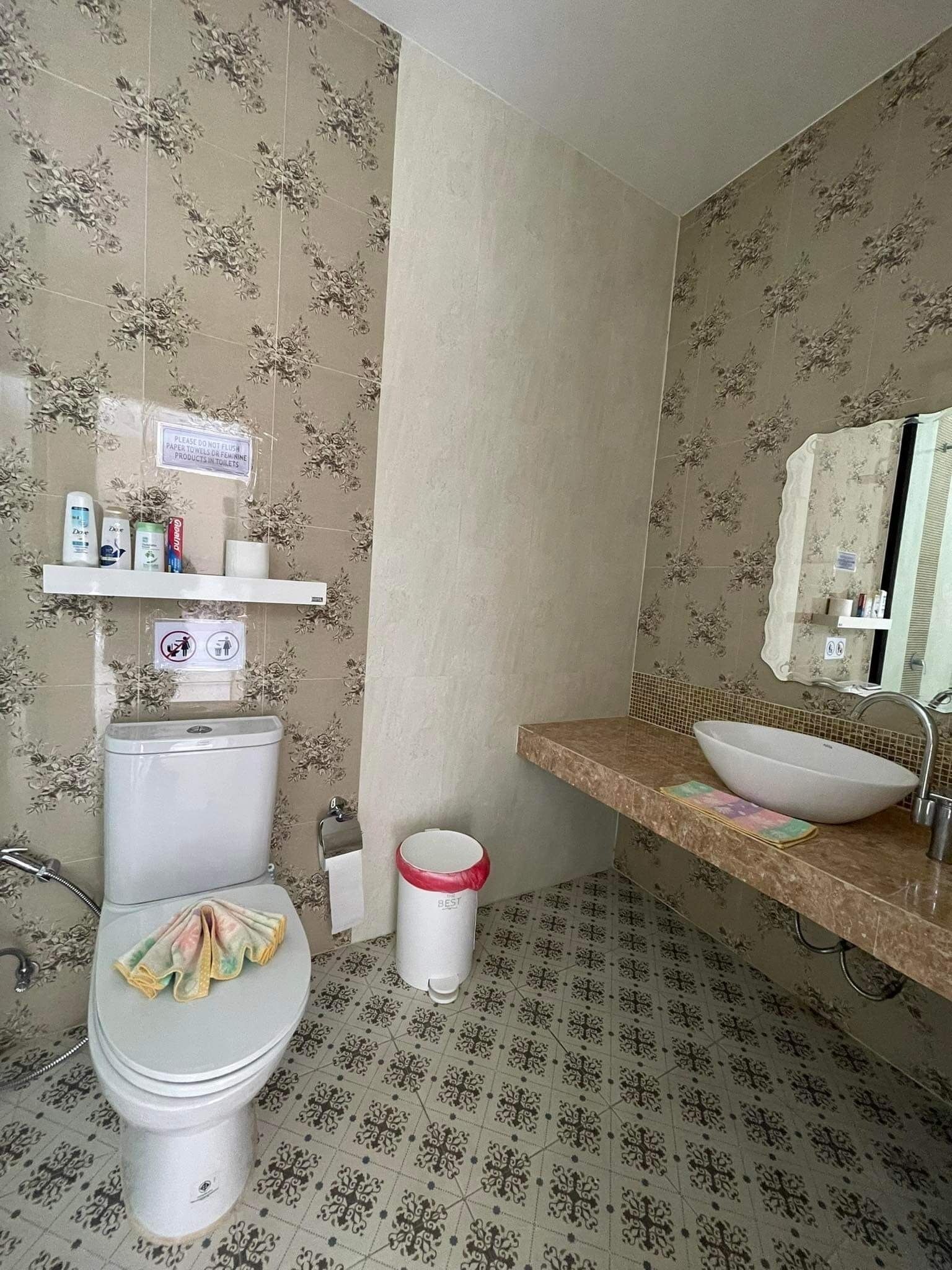A bathroom with a toilet, sink, and shower. There are toiletries on a shelf and a trash can.