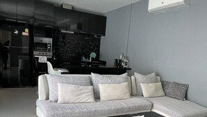 Uma sala de estar moderna com um sofá cinzento, uma mesa de centro preta e uma área de cozinha ao fundo.