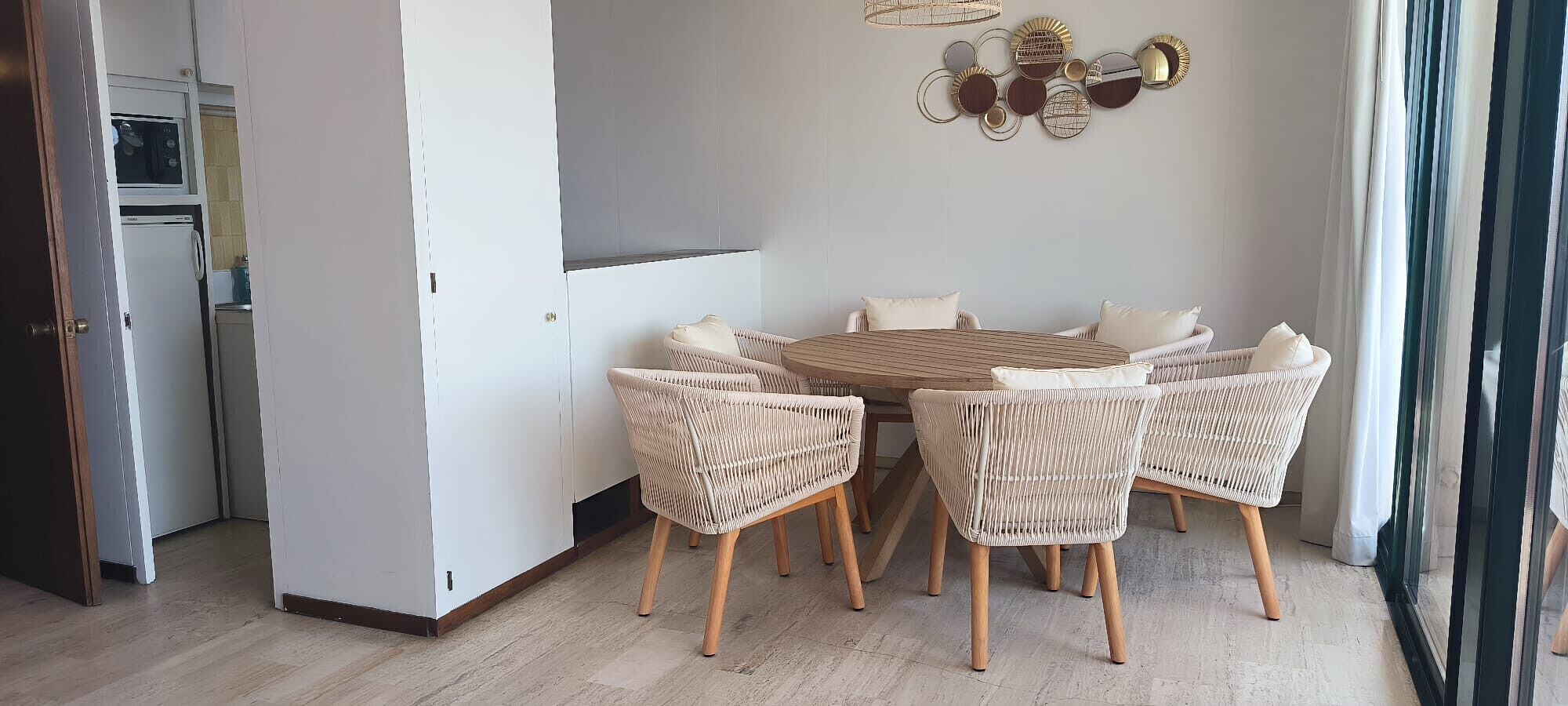 Een eetgedeelte met een ronde tafel en vier rieten stoelen, een keuken met een magnetron en koelkast, en een decoratief object aan de wand.