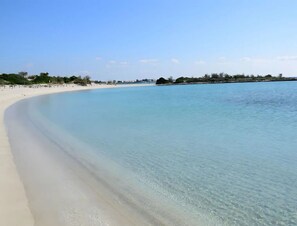 Ein Strand mit klarem Wasser, sandigem Ufer und einer entfernten Küstenlinie mit Vegetation und Gebäuden.