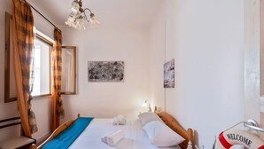 Una cama bien hecha con sábanas blancas y toallas enrolladas, un cabecero de madera, una manta azul, una lámpara de pared, un cuadro abstracto enmarcado y un cartel con la palabra 'WELCOME' en la pared.