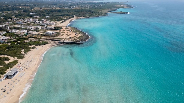 Vista aérea de una zona costera con playa, aguas turquesas cristalinas y una pequeña península con edificios y una estructura fortificada.