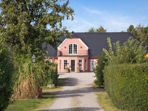 A two-story house with a dark roof and a pink facade, surrounded by greenery.