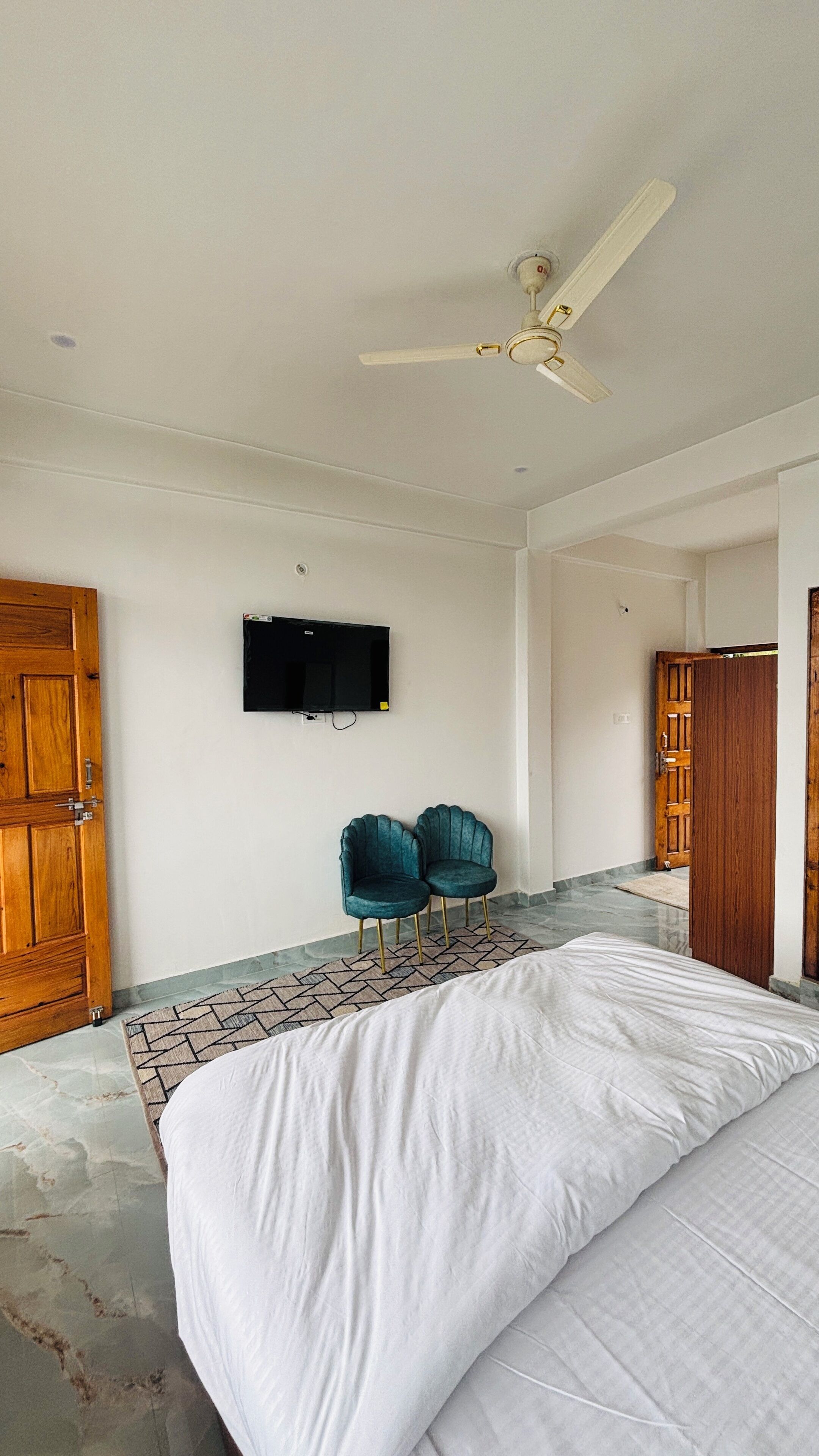 A bedroom with a bed, two chairs, a ceiling fan, a TV, and wooden doors.