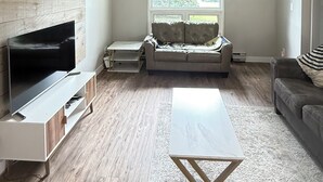 A spacious living room with a wooden wall, a large window, a sofa, a coffee table, and a TV on a stand.