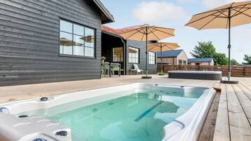 Un espace moderne aménagé à l’extérieur avec un bain à remous, une terrasse en bois et un coin salon couvert, offrant une vue sur une maison en arrière-plan.