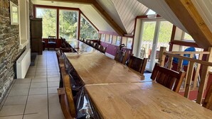 Une longue table en bois, entourée de chaises, se trouve dans une pièce aux murs de pierre et aux grandes fenêtres.