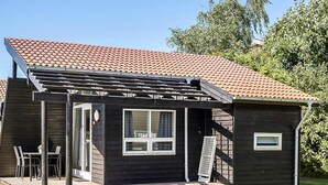 A small, dark wooden house with a tiled roof, a covered patio with chairs, and a window. - 6 Person Holiday Park Home in Bogense (Bogense)