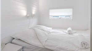 A white room with a bed, a small bedside table, and a window with blinds. - 6 Person Holiday Park Home in Bogense (Bogense)