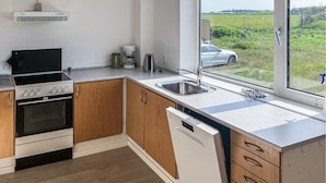 A modern kitchen with wooden cabinets, a built-in oven, a dishwasher, and a large window offering a view of a car and greenery outside. - 2 bedroom stunning home in Harboøre (Harboøre)
