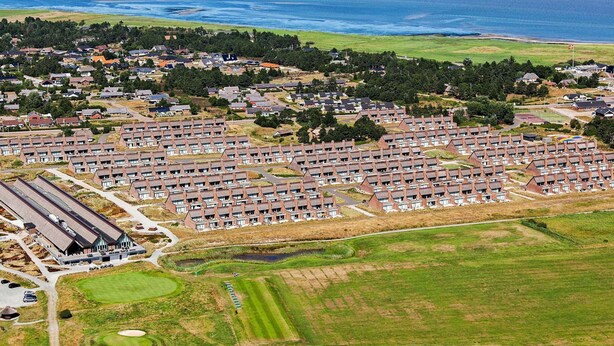 Een woongebied aan de kust met meerdere rijen huizen, een golfbaan en een groot waterlichaam op de achtergrond.