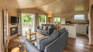 Ein gemütliches Blockhauss-Interieur mit Wohnbereich, einer Couch, Kamin und einer Küche mit Fensterblick.