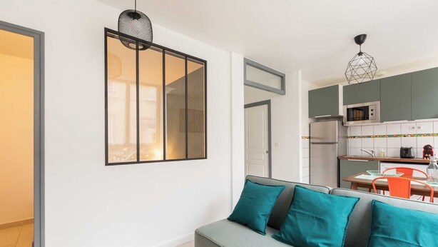 A modern living room with a grey sofa, teal cushions, a black-framed picture, and a kitchen in the background. - DIFY Berlioz - Charpennes (Villeurbanne)