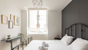 A neatly made bed with white linens and rolled towels, a bedside table with a plant, and a window with a view of buildings. - DIFY Berlioz - Charpennes (Villeurbanne)