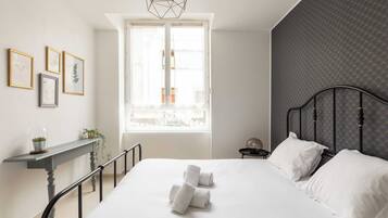A neatly made bed with white linens and rolled towels, a bedside table with a plant, and a window with a view of buildings.