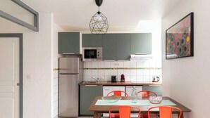 A compact kitchen with a microwave, oven, and refrigerator. There is a dining table with chairs and a hanging light fixture. - DIFY Berlioz - Charpennes (Villeurbanne)