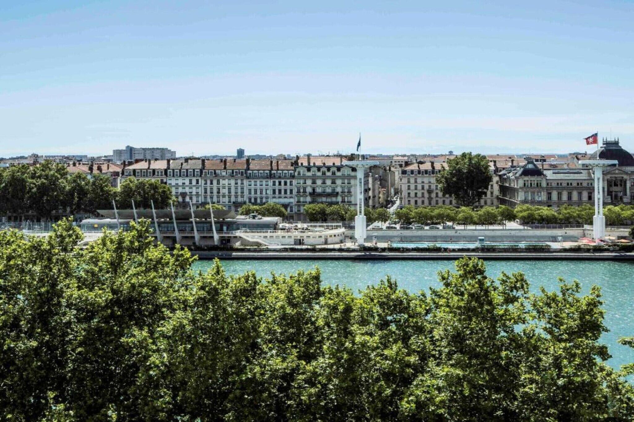 A waterfront cityscape with historic buildings, a modern bridge, and a flag on a tall structure.