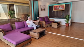 Eine moderne Lobby mit einem großen, violetten Ecksofa, einem Korbstuhl und einem kleinen Beistelltisch. Es befinden sich Blumentöpfe und Kunstwerke an den Wänden.