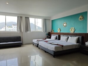 A hotel room with two beds, a sofa, and a large window with a view of buildings and mountains. - Sereneburi Patong Hotel By Sea Hospitality  Co Ltd (Patong)