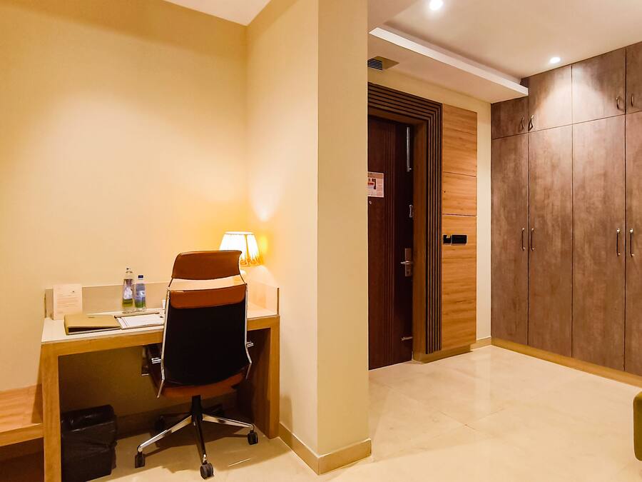Habitación de hotel con escritorio, silla, lámpara, armario y un reposapiés verde.