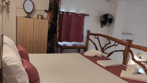 Une chambre à coucher avec un lit en bois, une chaise en bois, une armoire, un miroir, un ventilateur et un rideau.
