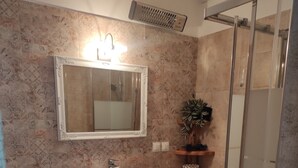 Une salle de bain avec un sol carrelé en marbre, un lavabo blanc doté d’un plan de travail en bois, un miroir au-dessus du lavabo, une douche avec une porte en verre et une plante en pot posée sur le plan de travail.