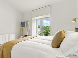 A bedroom with a large bed, a window with a view of greenery, a radiator, and a lamp. - The Loft @ the Warehouse (Clifden)