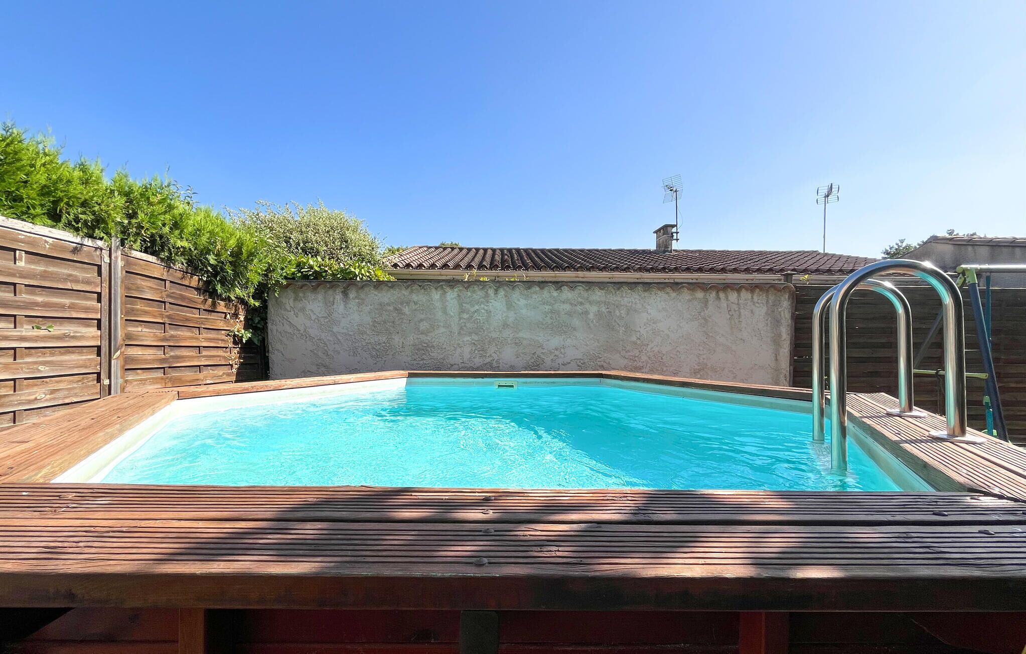 A swimming pool with a wooden deck and metal ladder, surrounded by a wooden fence and a tiled roof in the background.