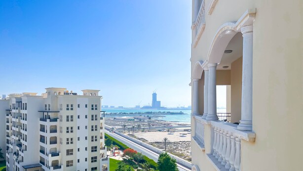 A high-rise building with multiple balconies overlooking a beach and marina. - Peaceful Lagoon View l 1BR in RAK With Balcony (Ras Al Khaimah)