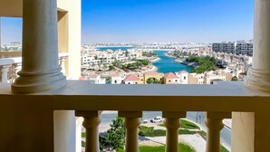A balcony with white columns overlooking a coastal cityscape with buildings, a river, and a clear blue sky. - Peaceful Lagoon View l 1BR in RAK With Balcony (Ras Al Khaimah)