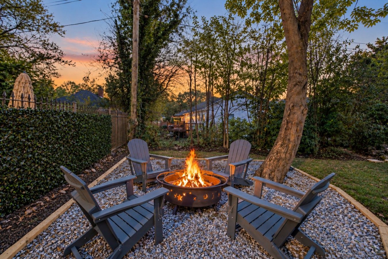 Ein Feuerkorb mit vier Adirondack-Stühlen, die ihn umgeben, auf einem Kiesplatz mit einer Hecke und Bäumen im Hintergrund.