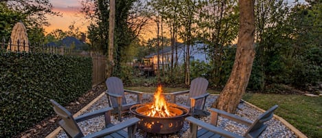 Ein Feuerkorb mit vier Adirondack-Stühlen, die ihn umgeben, auf einem Kiesplatz mit einer Hecke und Bäumen im Hintergrund.