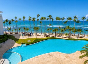 A pool with a curved edge, surrounded by a sandy area and palm trees, with a beach and ocean in the background. - Marbella | 120 M2• Beachfront • Pool • FastWiFi (Juan Dolio)