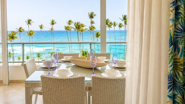 Mesa de comedor preparada para cuatro personas con sillas blancas, un bol grande y copas de vino, con vista a una playa con palmeras y el océano.
