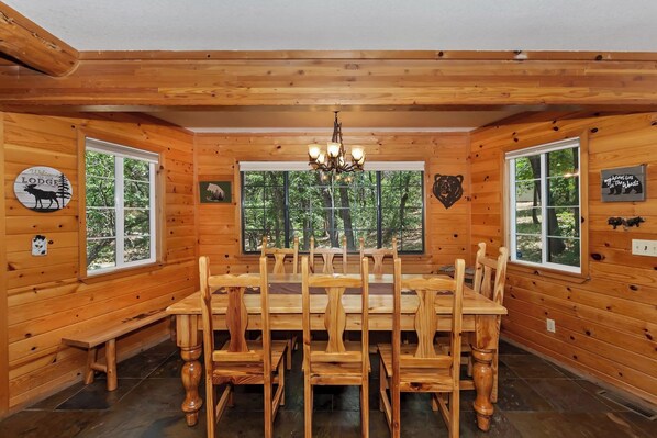 A wooden lodge dining room with a large table and chairs, large windows with forest views, and bear-themed decor.