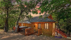 A wooden cabin with a hot tub, a deck, and a swing set, surrounded by trees. - Single-Story! Pet Friendly! Hot Tub! Pool Table! (Big Bear City)