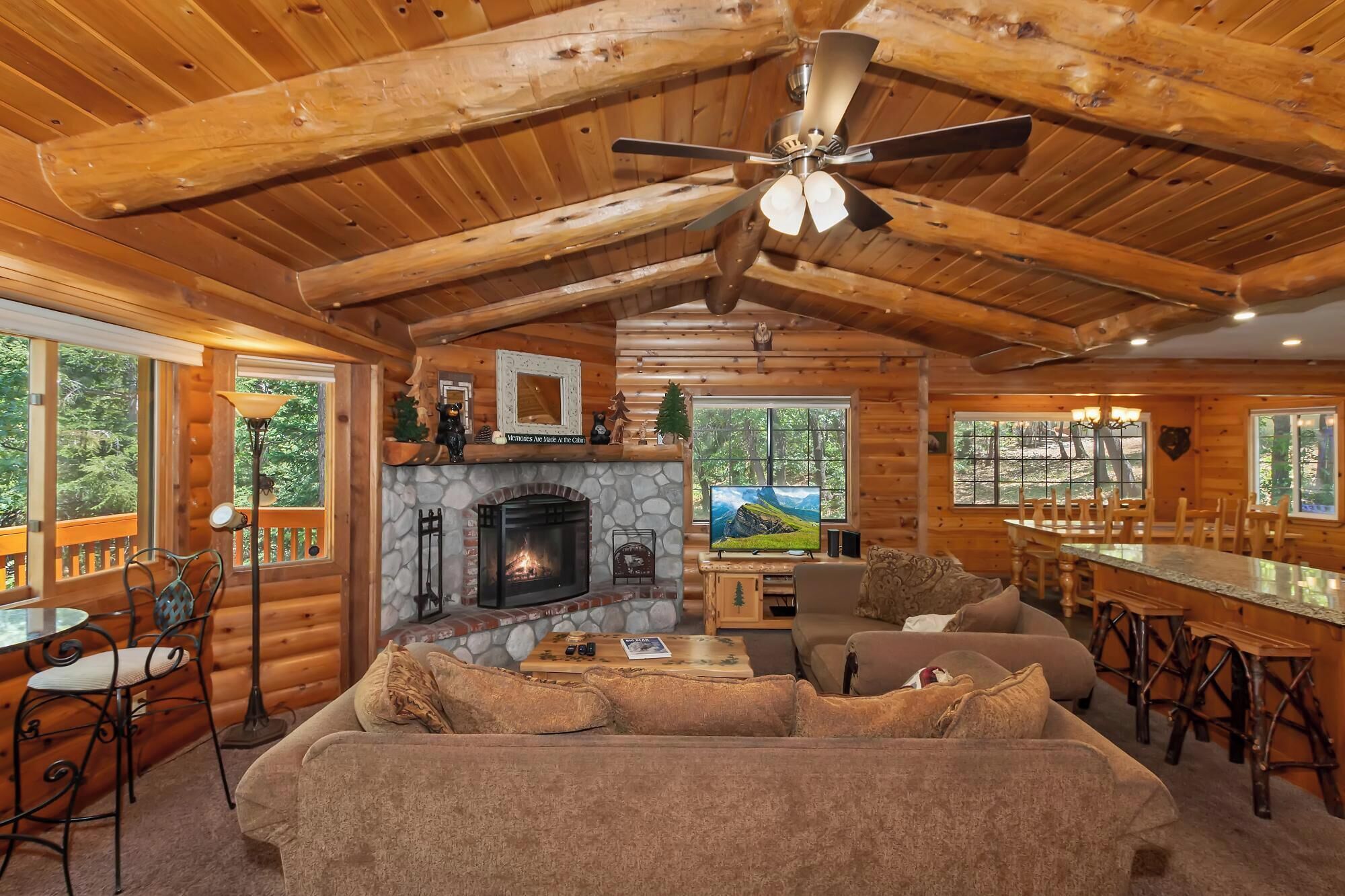 L’intérieur d’une cabane en rondins est doté d’une cheminée, d’un ventilateur de plafond, d’un coin repas et de grandes fenêtres offrant une vue sur la forêt.