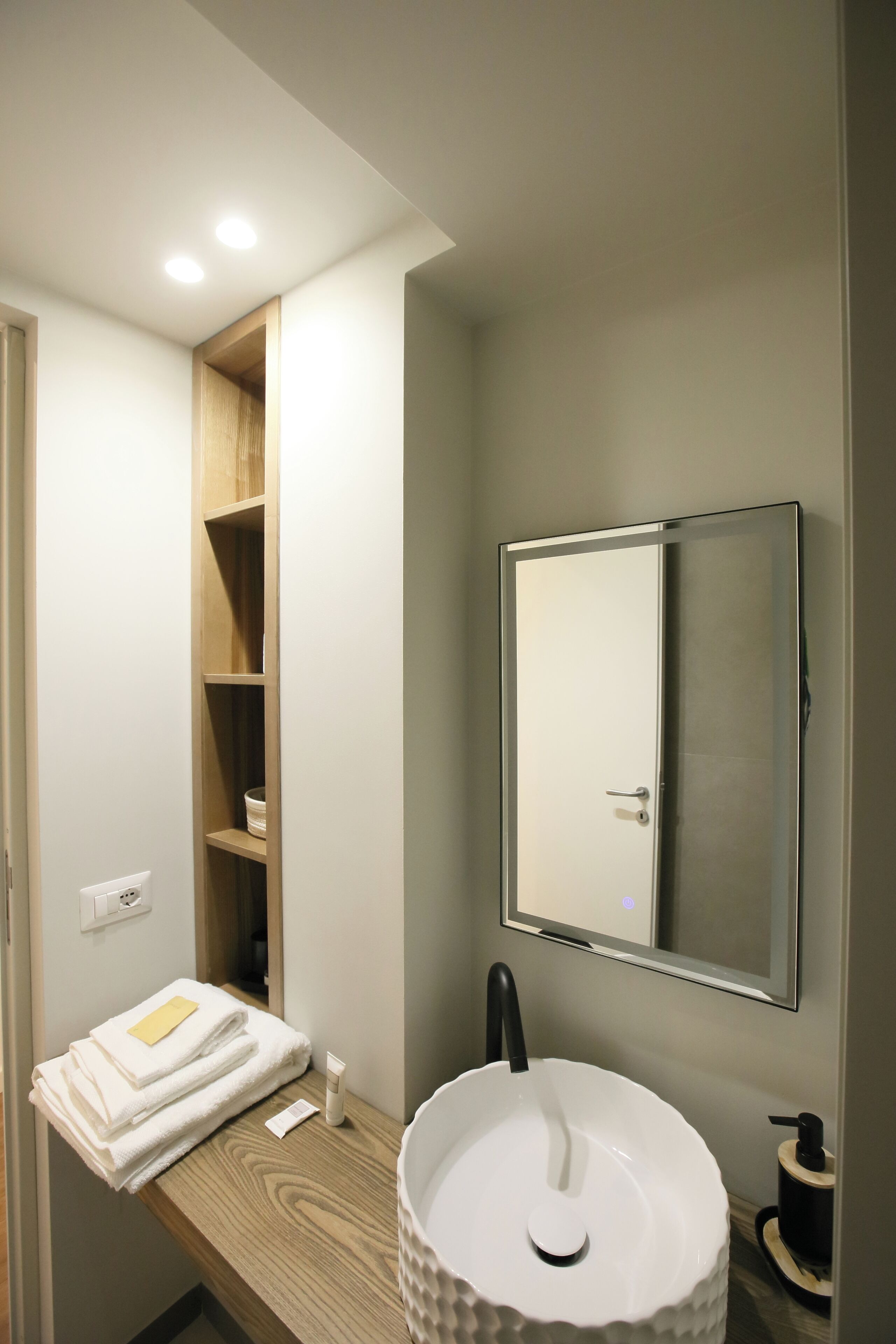 Une salle de bain avec un lavabo blanc, un miroir et une étagère en bois où sont posées des serviettes.