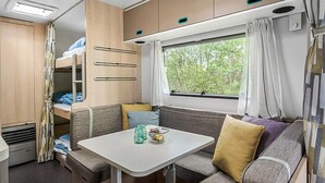 Een compact camperinterieur met een bunkebed, een eettafel en een raam met uitzicht op groen.