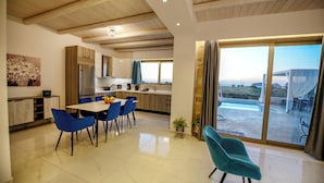 A modern kitchen with wooden cabinets, a dining area with blue chairs, and a large sliding glass door leading to an outdoor patio. - Koukis Villa With Sea View (Kissamos)